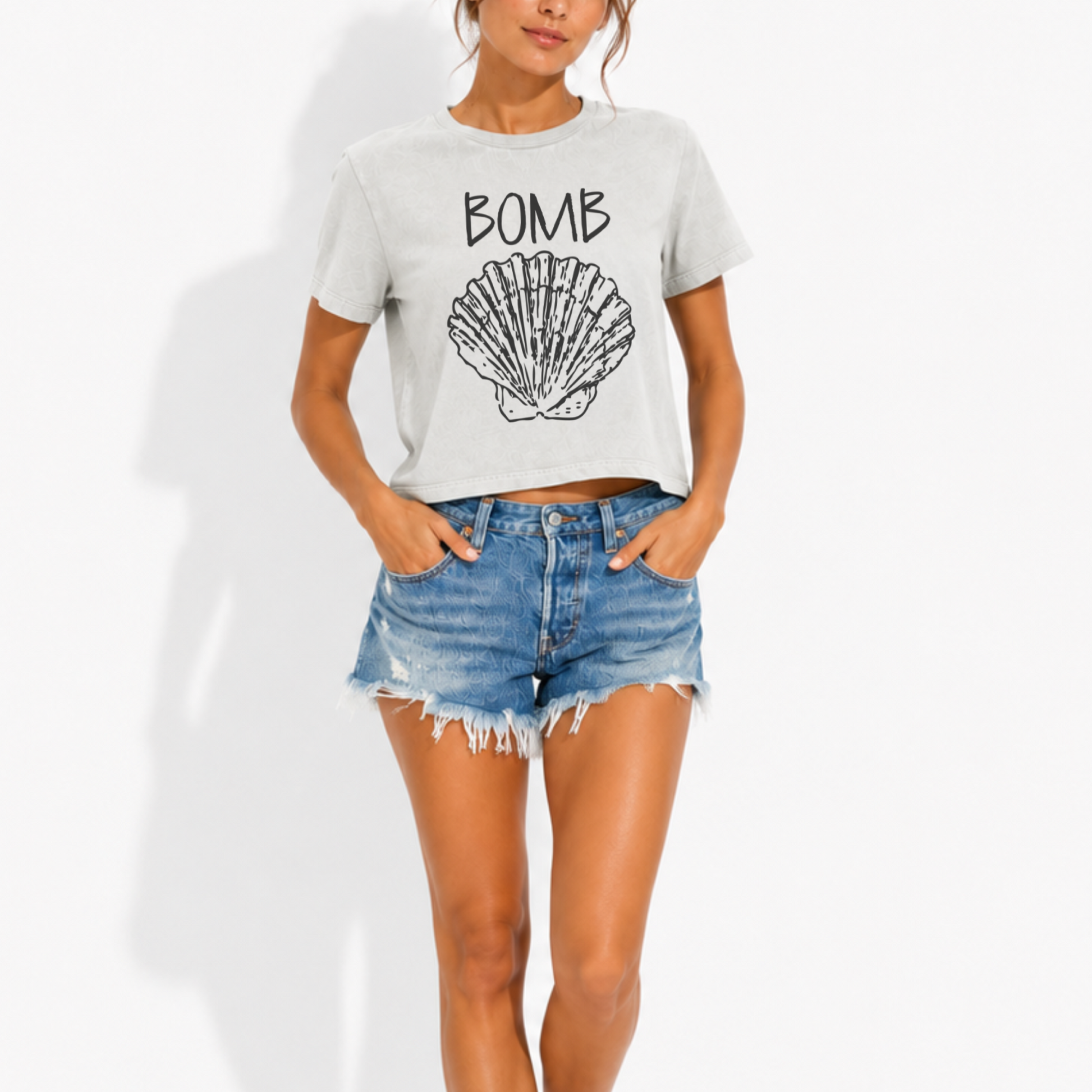 Woman wearing a washed garment-dyed t-shirt featuring the phrase "Bomb Shell" with a detailed seashell graphic, creating a fun vintage-inspired beach look.