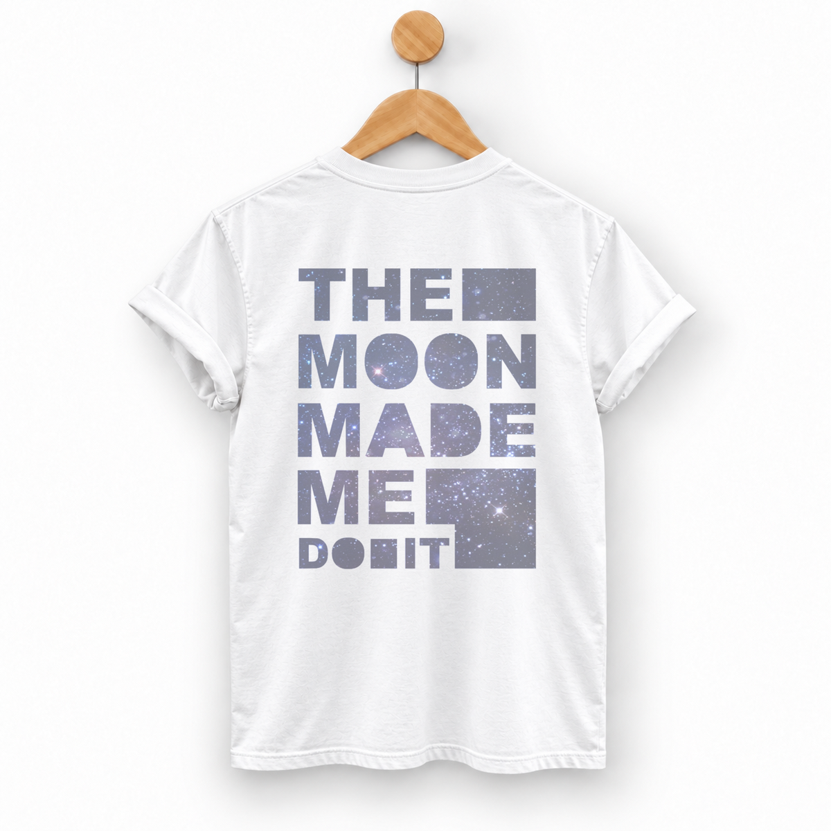 A white short-sleeve T-shirt hanging on a wooden hanger against a light gray background. The back of the shirt features a large graphic text print reading 'THE MOON MADE ME DO IT' in a dusty purple-gray galaxy pattern.