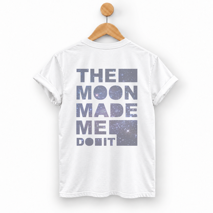 A white short-sleeve T-shirt hanging on a wooden hanger against a light gray background. The back of the shirt features a large graphic text print reading 'THE MOON MADE ME DO IT' in a dusty purple-gray galaxy pattern.