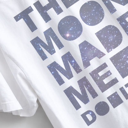 White short-sleeve T-shirt with large dark blue text reading 'THE MOON MADE ME DO IT' filled with a starry galaxy pattern, shown in a close-up studio-style shot. The Moon Made Me Do It boyfriend tee featuring a celestial astrology graphic on a garment dyed unisex T-shirt with relaxed sizing and a slightly oversized fit for women.
