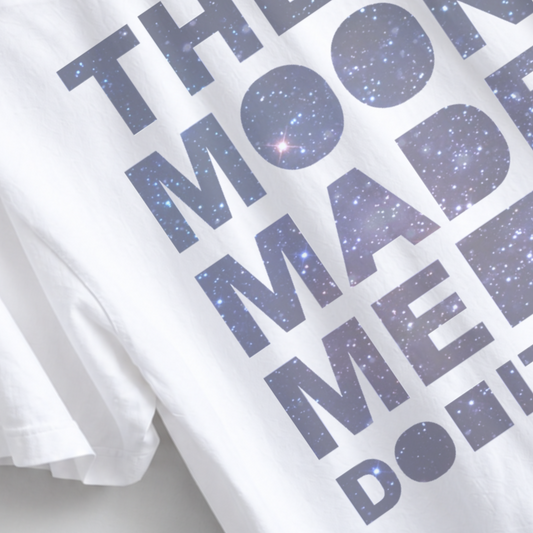 White short-sleeve T-shirt with large dark blue text reading 'THE MOON MADE ME DO IT' filled with a starry galaxy pattern, shown in a close-up studio-style shot. The Moon Made Me Do It boyfriend tee featuring a celestial astrology graphic on a garment dyed unisex T-shirt with relaxed sizing and a slightly oversized fit for women.