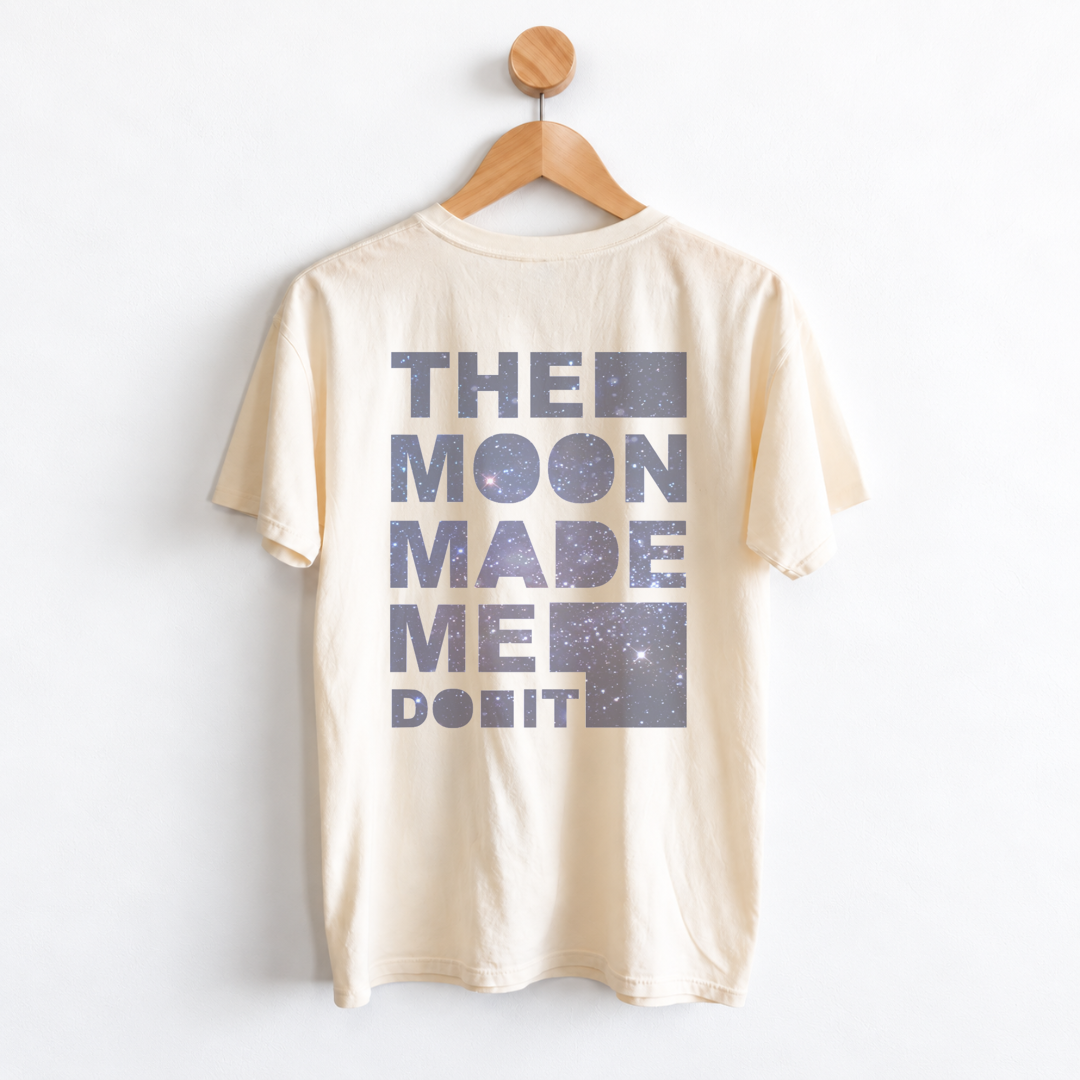 A cream-colored t-shirt hanging on a wooden hanger against a white wall. The back of the shirt features a large graphic print in a dusty blue-purple color that reads 'THE MOON MADE ME DO IT' with starry galaxy patterns inside the letters and block shapes.